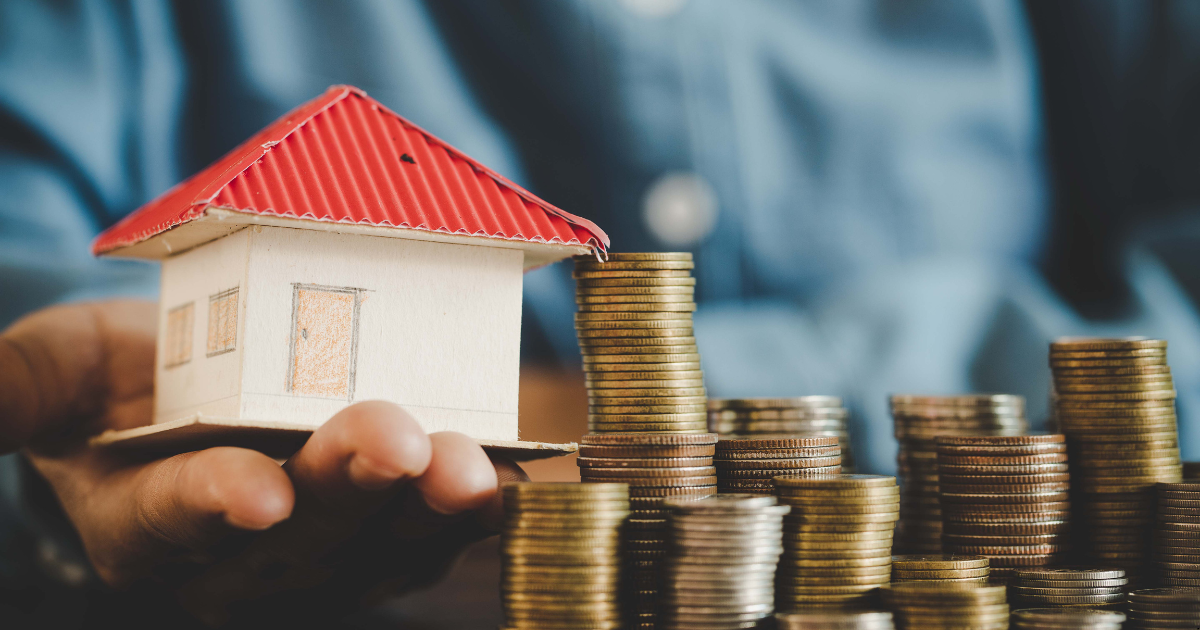 a person holding a dummy house with a lot of coins on the table showing the cost of owning the house is more than the property rental income which is basically negative geared property